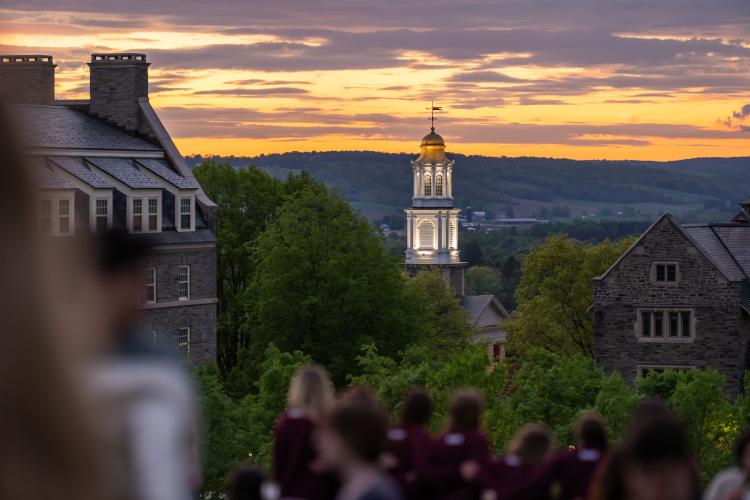 Illuminated bell tower of a university building at sunset, framed by trees and stone campus buildings, with students in the foreground looking toward the horizon.